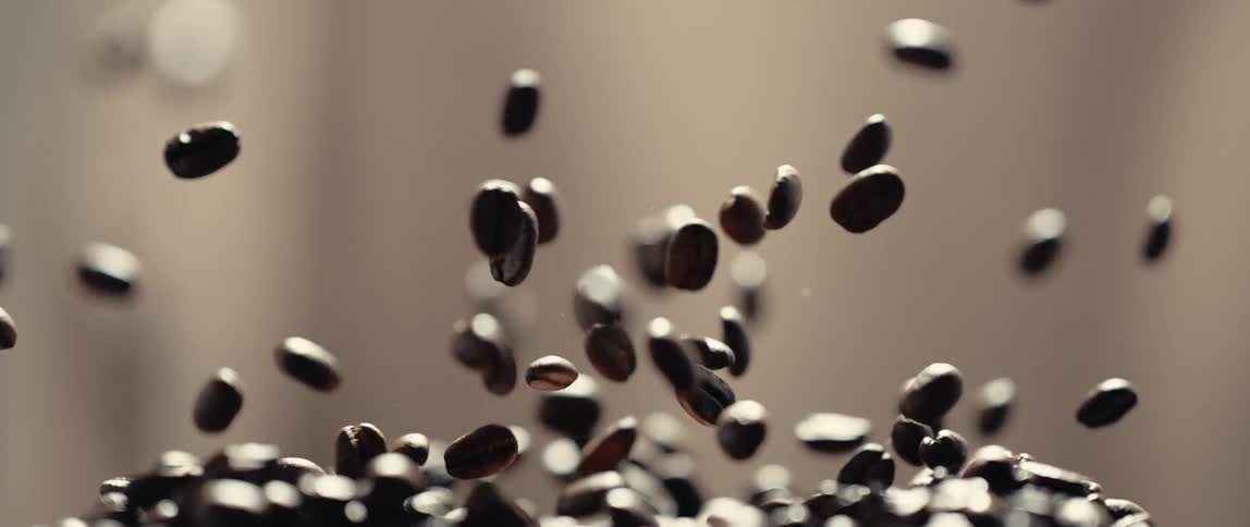 The clip is a slowmotion closeup shot of dark freshly roasted coffee beans bouncing and raining down onto a pile of other beans at the bottom of the framethe background is uniformly light and softfocus emphasizing the texture and motion of the i