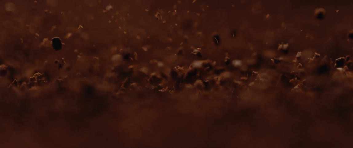 An extreme closeup shot captures dark brown irregularly shaped granules resembling coffee grounds falling and swirling against a dark brown blurred background