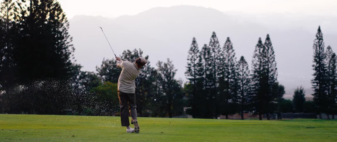 A man swings a golf club on a green course with trees and mountains at sunset