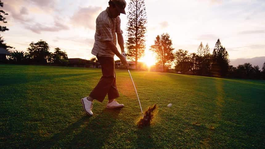 Golfer stands with his club in the air and swings to hit the golf ball as the sun shines on the horizon