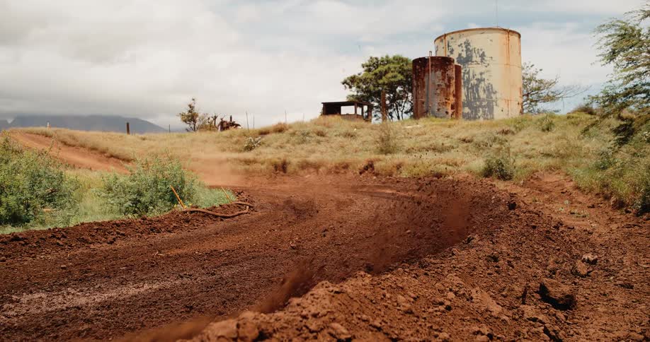 Rider rounds bend on dirt track surrounded by vegetation and rusted industrial tanks