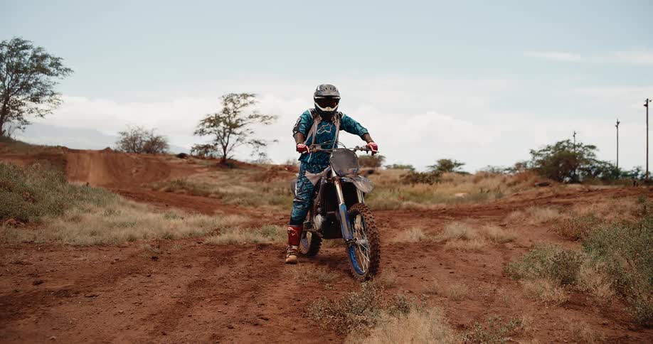 Motocross rider rests on bike along dry trail surrounded by sparse vegetation under clear sky