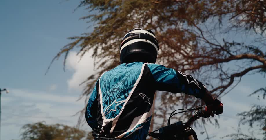 A rider wearing a full face helmet and blue patterned motocross jersey looks back before riding off screen on a sunny day