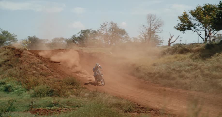 A motocross rider soars over a hill at midday landing firmly before continuing along the dirt bike path