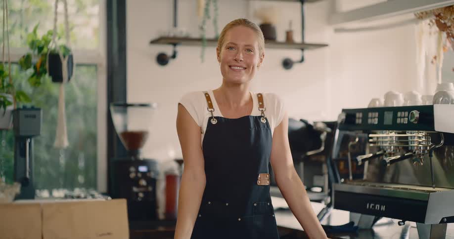 Smiling barista in black apron serves behind counter with espresso machine and hanging plants