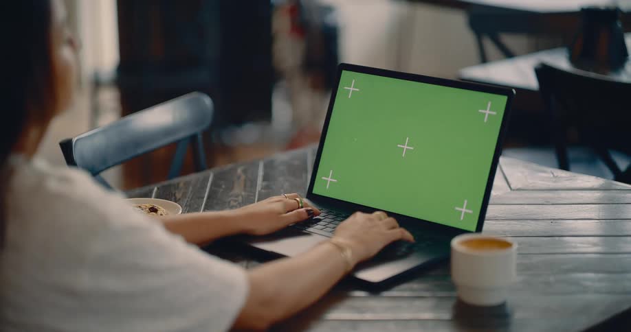Woman sits at a table typing on a computer with a green screen as she reaches for a cup of coffee