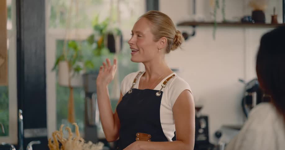 This video features a friendly barista in a white shirt and dark apron standing behind the counter of a bright cafe she is smiling and conversing with a customer who is visible over her right shoulder