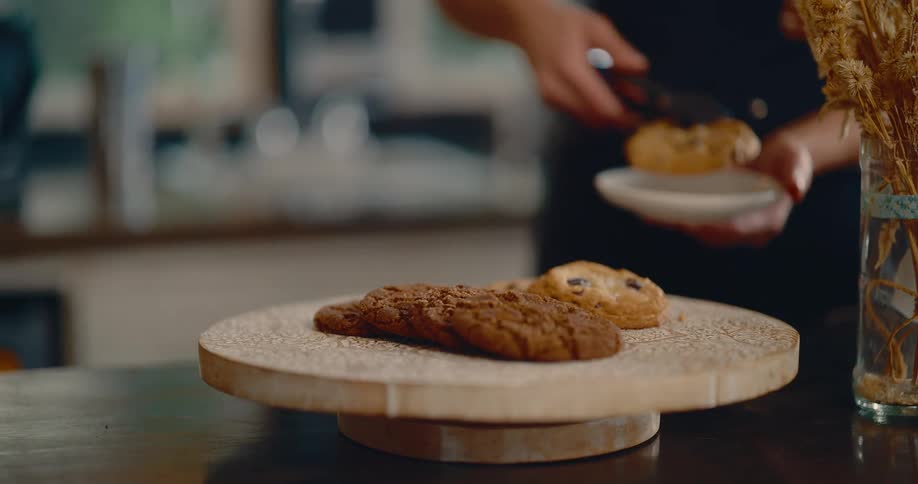 Assorted cookies rest on wooden board as hand with tongs serves portion onto white plate