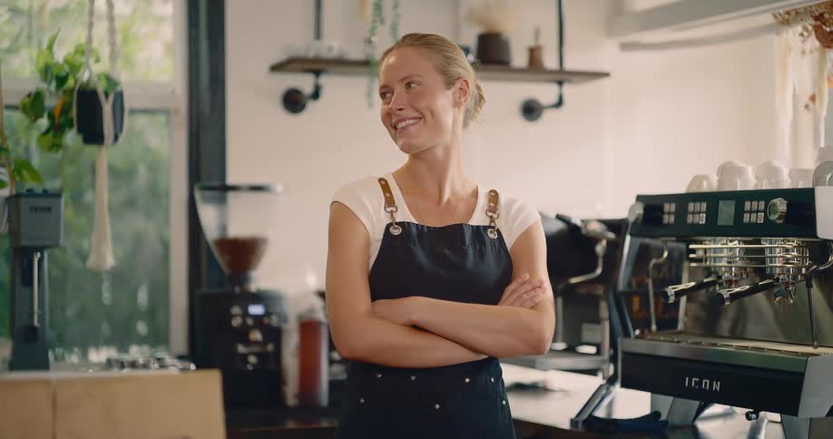 A smiling woman wearing a white shirt and a black apron stands in the middle of a brightly lit coffee shop with her arms crossed behind her is a professional espresso machine and coffeemaking equipment highlighting her role as a barista or shop owner