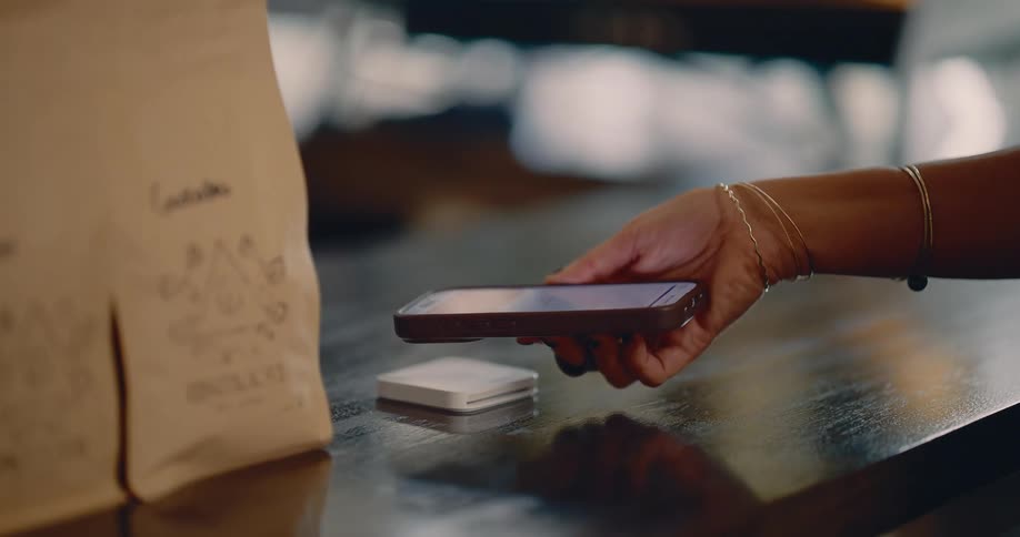 A closeup shot shows a persons hand holding a smartphone near a small white square payment terminal on a dark wooden counter the person is performing a contactless mobile payment like apple pay or google pay for an item that is in a brown paper bag