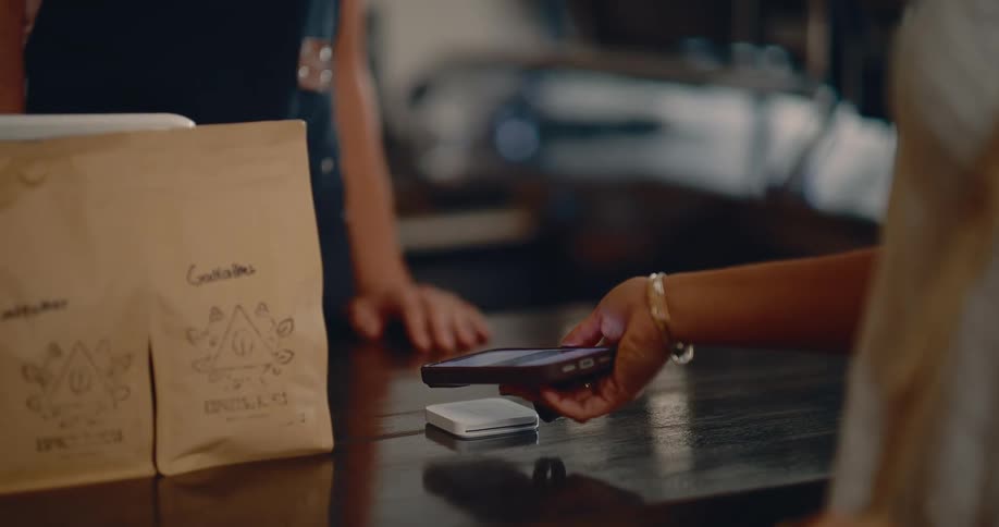 A persons hand holding a cell phone and bringing it close to a small payment terminal on a table
