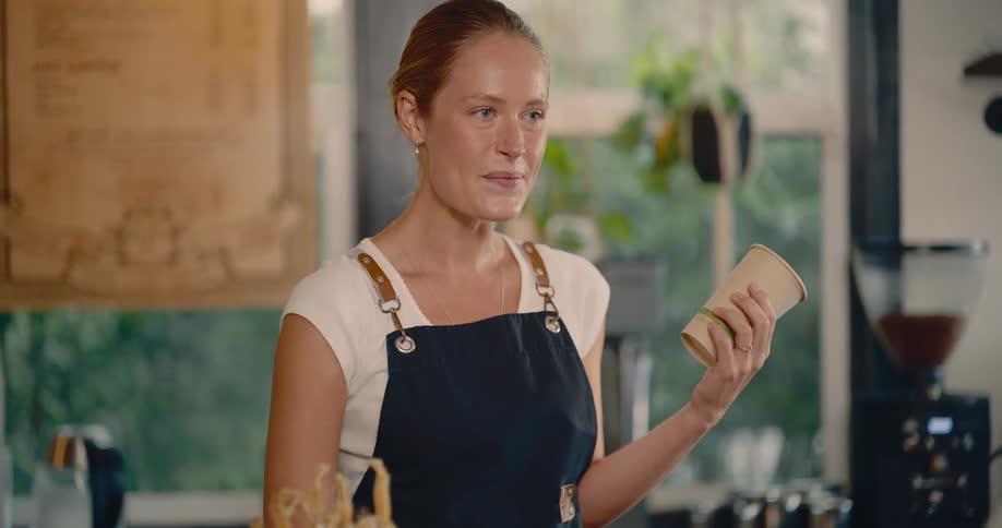 A smiling woman wearing a dark apron writes a name on a blank cardboard coffee cup in a brightly lit coffee shop setting