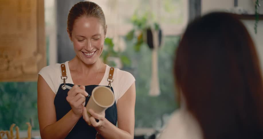A close view shows a smiling woman barista wearing an apron talking to a customer while using a pen to write on a disposable coffee cup