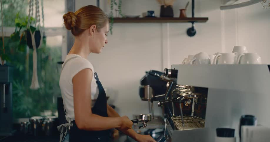 A woman with her hair pulled back wearing a white shirt and a black apron is shown working as a barista behind a counter she is focused on wiping down the portafilter of a professional espresso machine in a welllit coffee shop setting