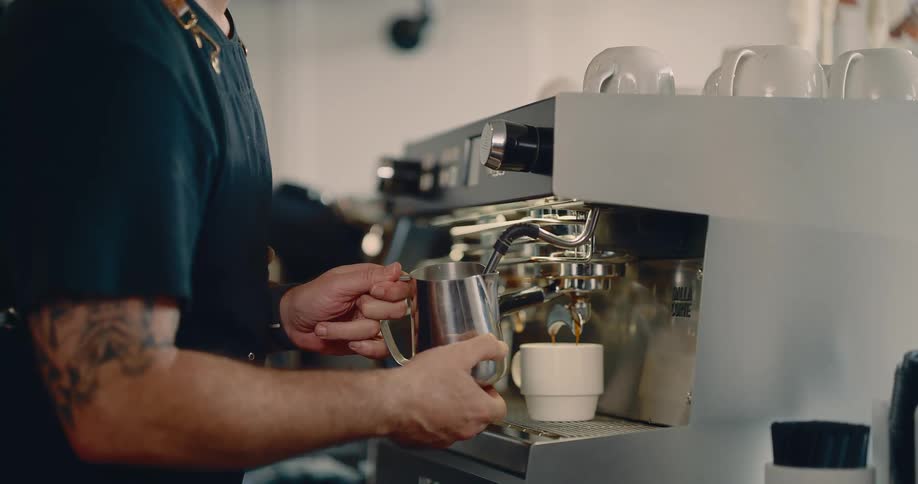 A closeup shot of a barista shows him holding a metal pitcher under the steam wand while simultaneously watching espresso pour into a white cup from the machines portafilter