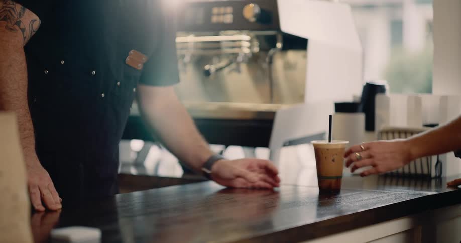 A close view shows a barista handing an iced coffee beverage to a customer across a dark wooden counter with a professional espresso machine visible