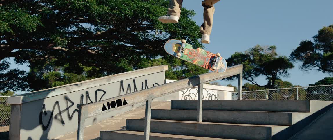 This video captures a skateboarder midair performing a trick over a set of concrete stairs and a metal railing in a skate parkthe skateboarder is wearing light brown pants and white shoes and the skateboard features a colorful graphic design on its
