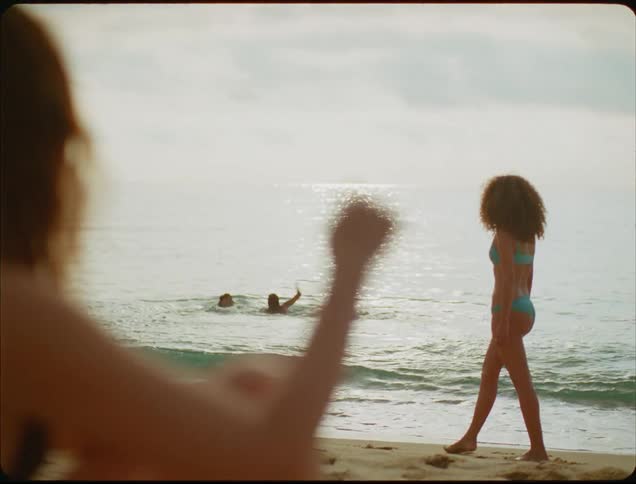 A woman with curly hair wearing a teal bikini stands on a sandy beach and gestures while looking toward the camera which is positioned behind the shoulder of another person in the foreground as two other individuals play in the waves behind her