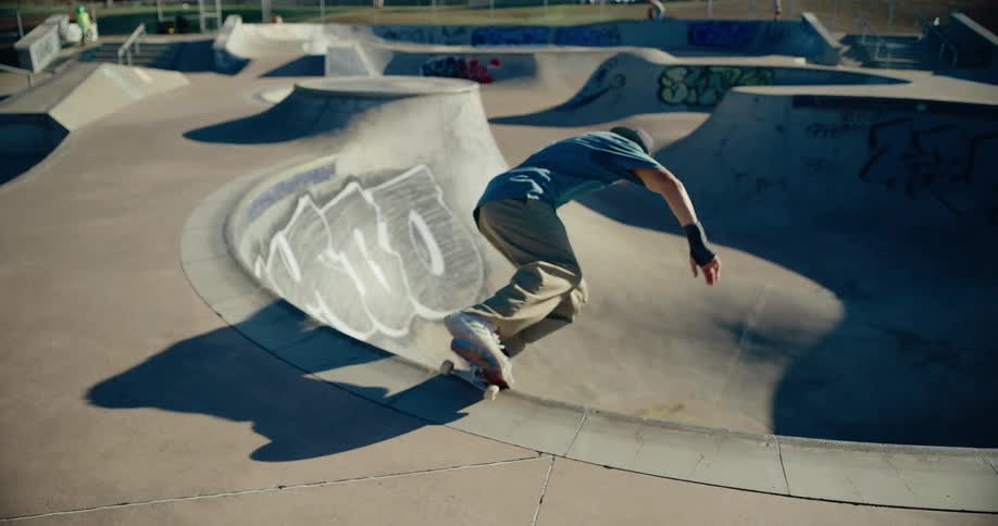 The video shows a skateboarder riding in a concrete skate park which features various bowls and rampsthe skater performs a trick riding up the curved wall of a bowl briefly hanging in the air and then coming back down to continue riding