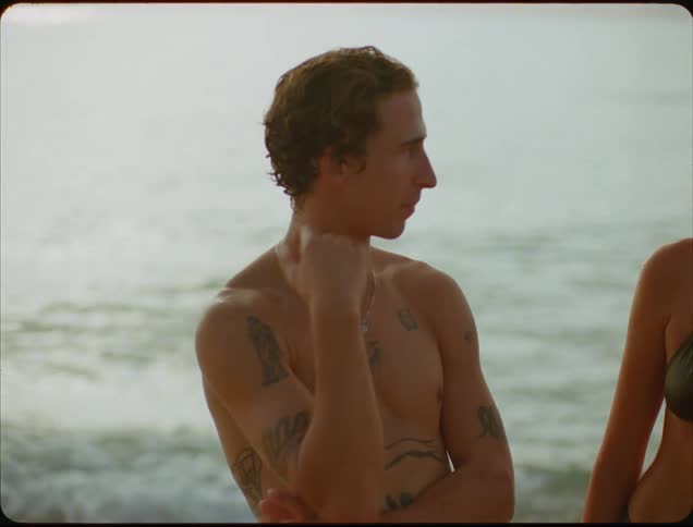 A shirtless young man with tattoos stands on the beach at midday beside a young woman in a bikini the two engaged in conversation as ocean waves crash onto the shore in the background