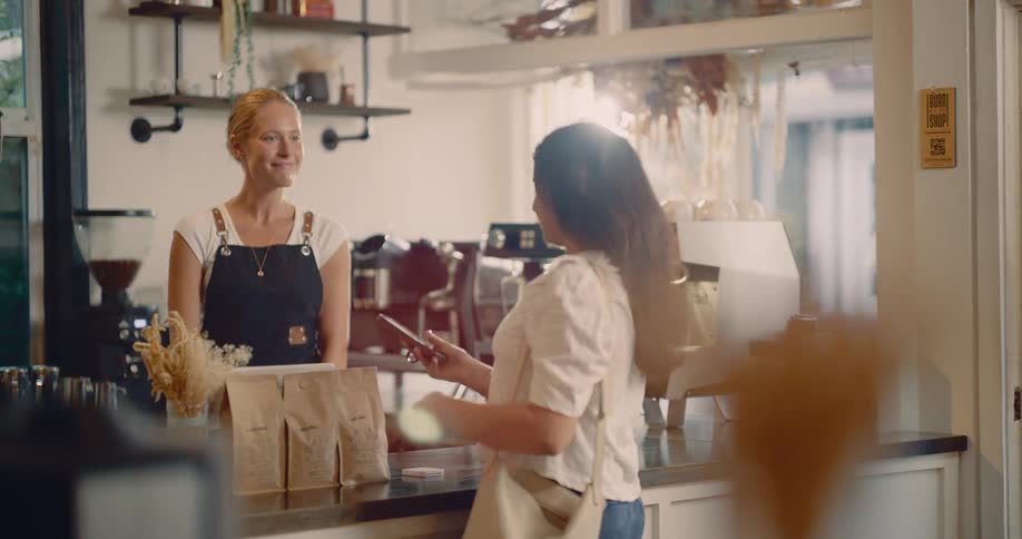 A customer is seen finishing a phone call as she enters a bright coffee shop where a friendly barista waits behind the counter the customer then prepares to pay using her smartphone indicating a modern casual transaction