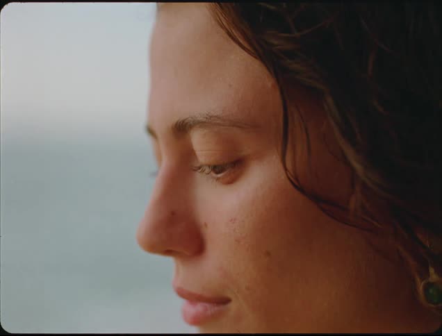 A close view shows a young womans profile with dark hair and a serious expression looking out toward the blurry ocean on an overcast day