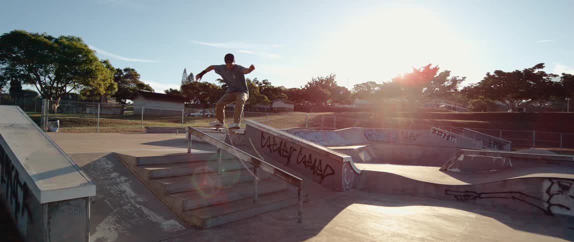 A skateboarder performs a grind down a handrail at a sunny skatepark
