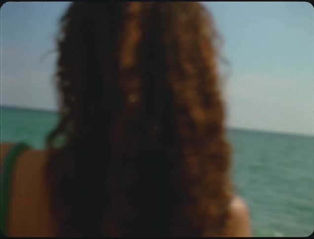 Three people in swimwear walk on sand near the sea under partly cloudy sky