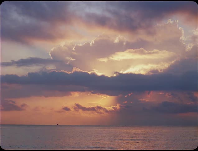 Clouds over the water in a colorful sky as sunlight reflects on the water and a ship sails in the distance