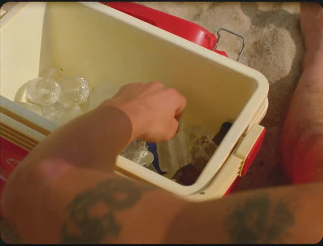 A tattooed man is sitting in sand as he grabs a bottle out of an ice cooler