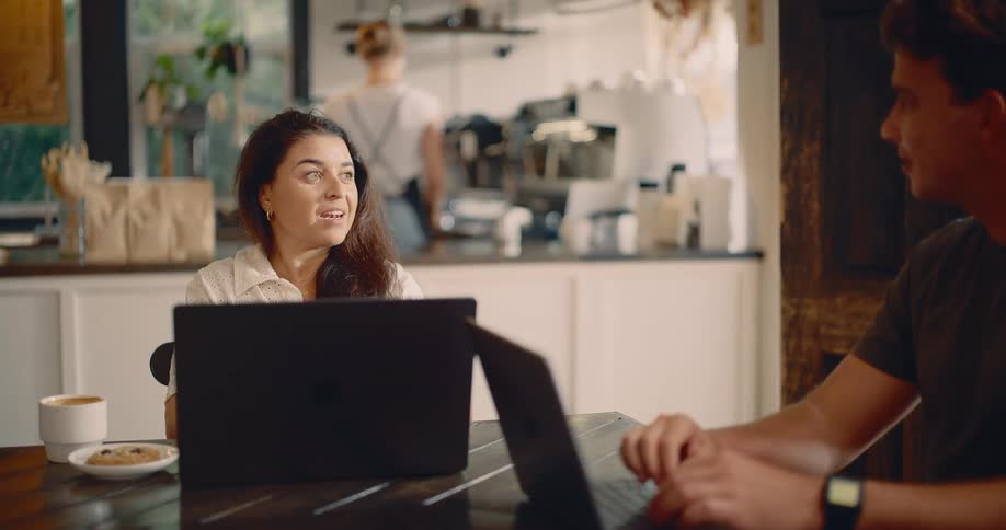 A man and woman are at a table with their laptops and coffee as a barista works in the background of a coffee shop