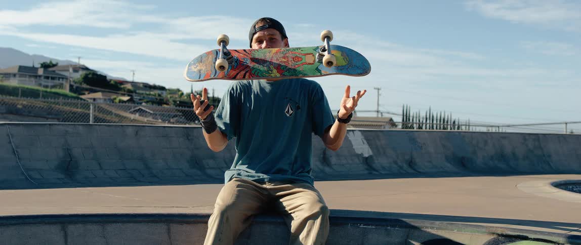 A close view shows a smiling man wearing a baseball cap sitting in a skatepark holding a colorful skateboard in front of his face