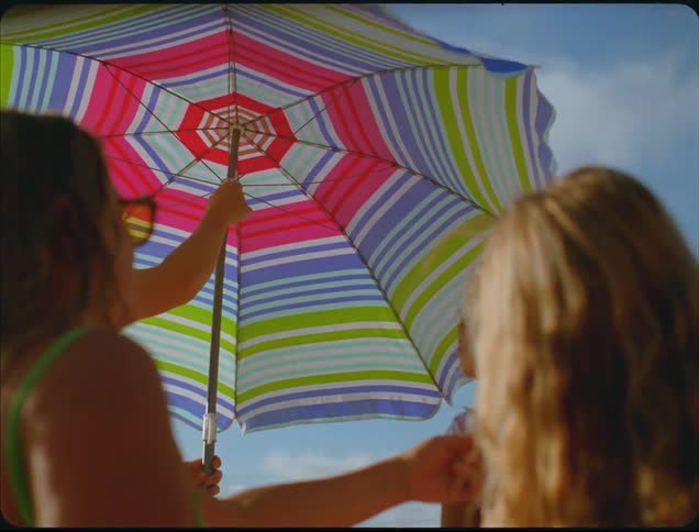 Person adjusts a striped beach umbrella while talking to someone nearby