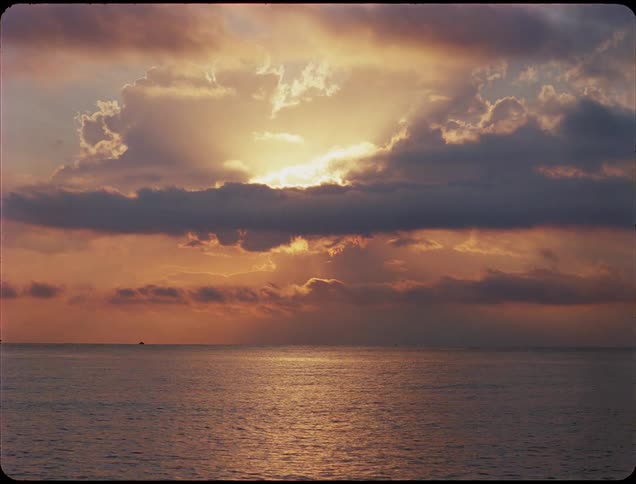 A sunset over the ocean where the sun bursts through a layer of dark clouds casting a strong golden reflection on the rippling water below
