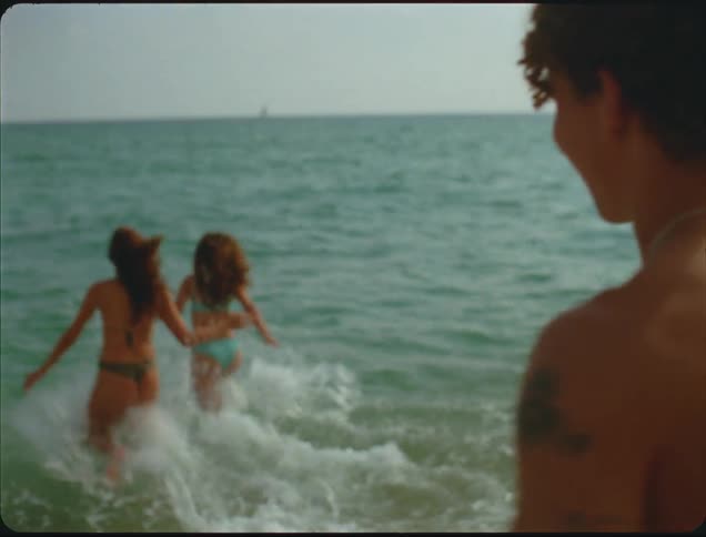 A close view shows two women in swimsuits running into the ocean waves while a man stands watching them from the sand on a warm day