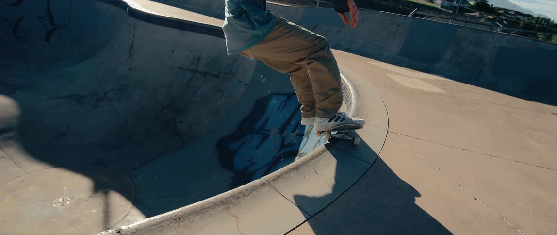 A close view shows a man performing a difficult skateboard maneuver on the rim of a large concrete bowl at an outdoor skatepark