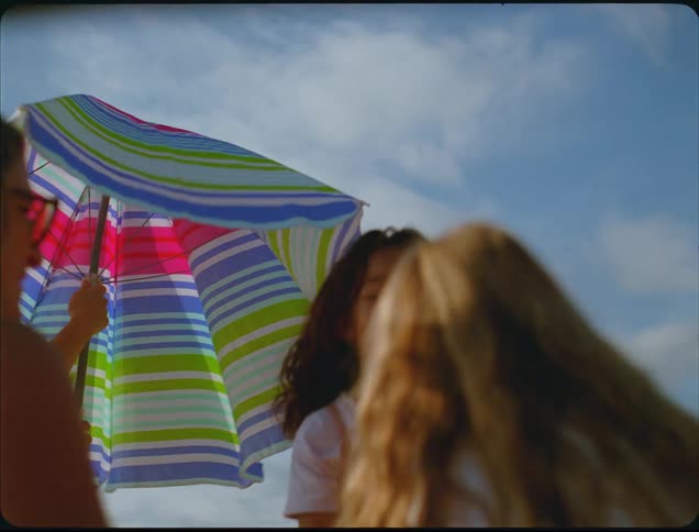 A close view shows a person adjusting a colorful striped umbrella on a sunny day with a blue sky surrounded by friends