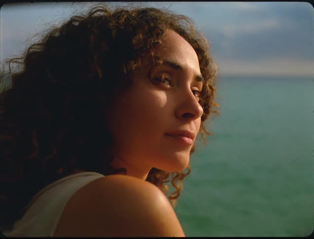 A close side profile of a young woman with long curly brown hair looking out over the sparkling green ocean under a bright sky