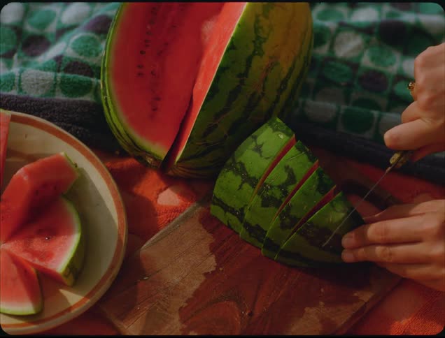 A person uses a knife to cut large triangular slices from a whole watermelon on a wooden cutting board with other precut slices of the fruit resting on a plate nearby all situated on a patterned towel