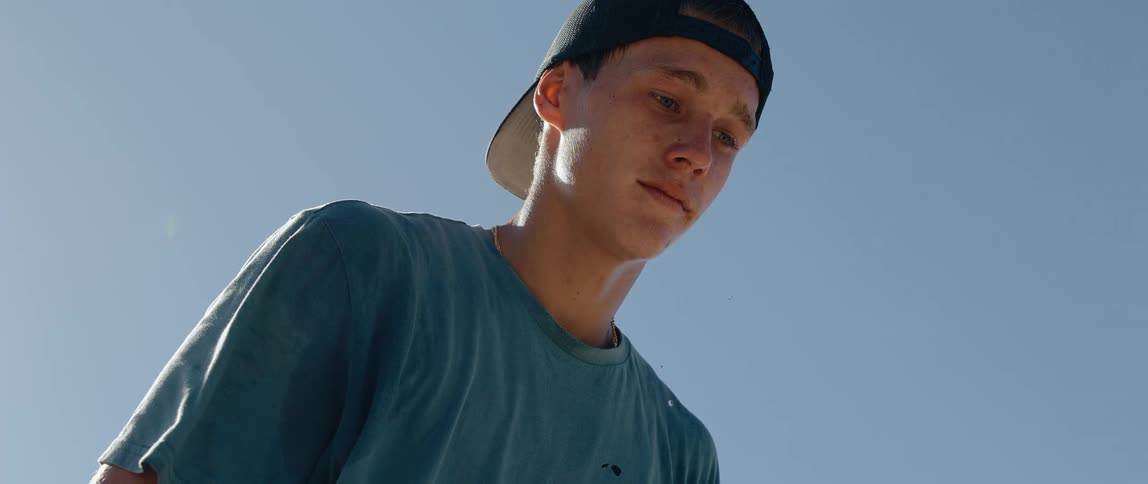 A close view shows a young man wearing a dark cap and blue shirt looking down intently with drops of water running down his face