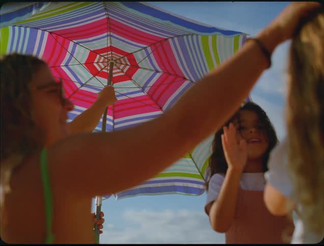 A closeup shot captures one women and two girls standing together on a sunny day framed by a large colorful striped beach umbrella held up against the blue sky