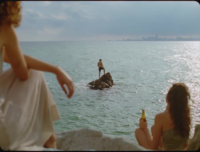 A man is on a rock in the water as two women sit on shore and drink