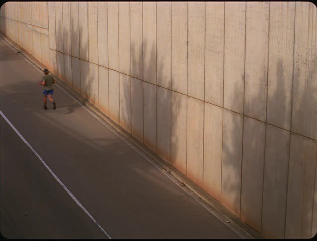 Person jogs beside concrete wall with tree shadows cast by sunlight