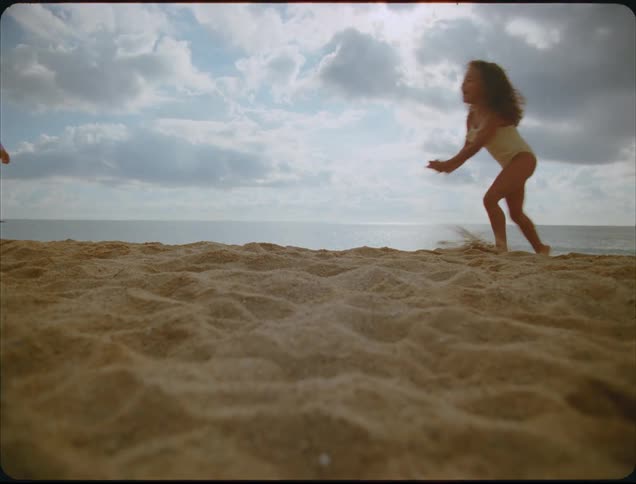 A close view shows a young girl running and jumping on a textured sand dune against a bright cloudy sky above the dark ocean