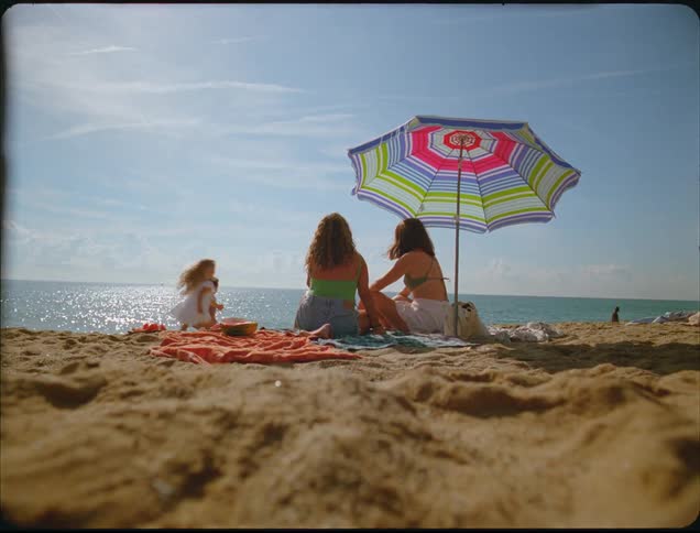 Two young women sit on the beach sand under a brightly colored umbrella by the shining ocean while little girls run and play around them