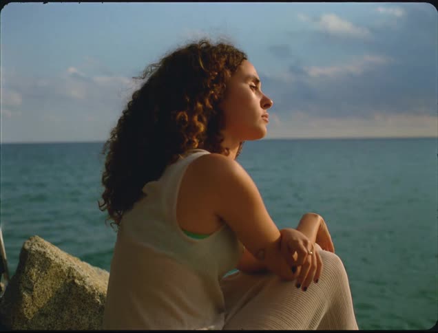 A young woman with curly brown hair sits peacefully on coastal rocks near the deep blue ocean under a pale blue sky
