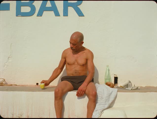 A shirtless elderly man seated in an outdoors setting drinking from a yellow mug