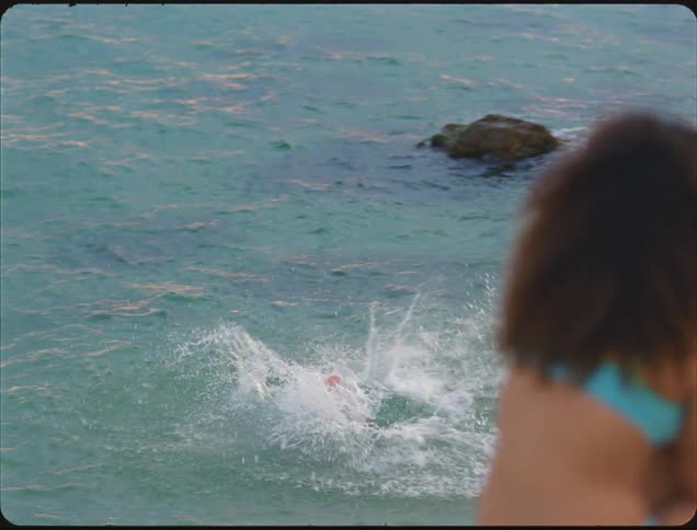 A close view shows a man running down a rocky sandy beach and diving into the clear turquoise ocean water