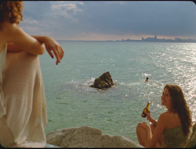 A man dives into the ocean from a rock as two women sit on the cliffs in the foreground drinking beer with a distant city skyline visible across the sparkling water