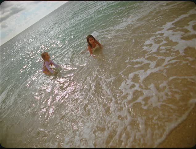 A close view shows two young girls playing excitedly in the small foaming waves washing onto the wet sand of a sunny beach